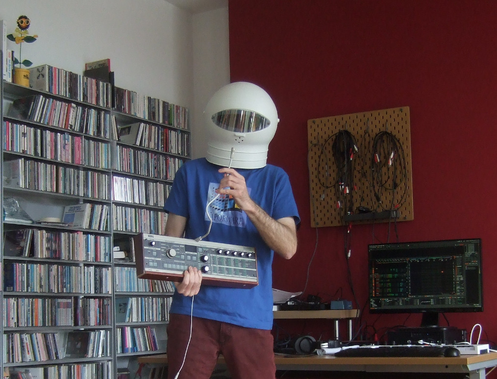 Marinus Nullbyte, wearing a space helmet while holding a small synthesizer which has a microphone attached to it