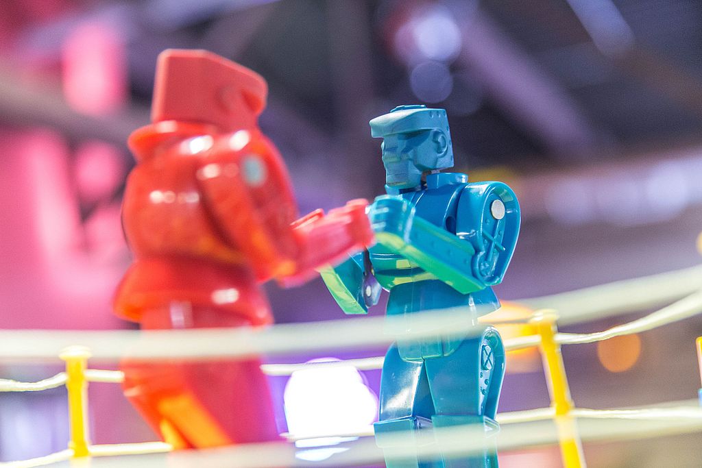 Photo of a red toy robot and a similar looking blue toy robot in a boxing ring. Both robots face each other in a threatening stance.