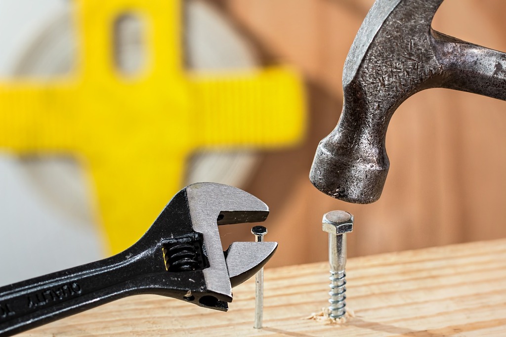 Photograph that shows a hammer that is used to smash a screw into a piece of wood. On the left is a nail that is partially pushed into the same piece of wood, with an adjustable wrench immediately next to it.